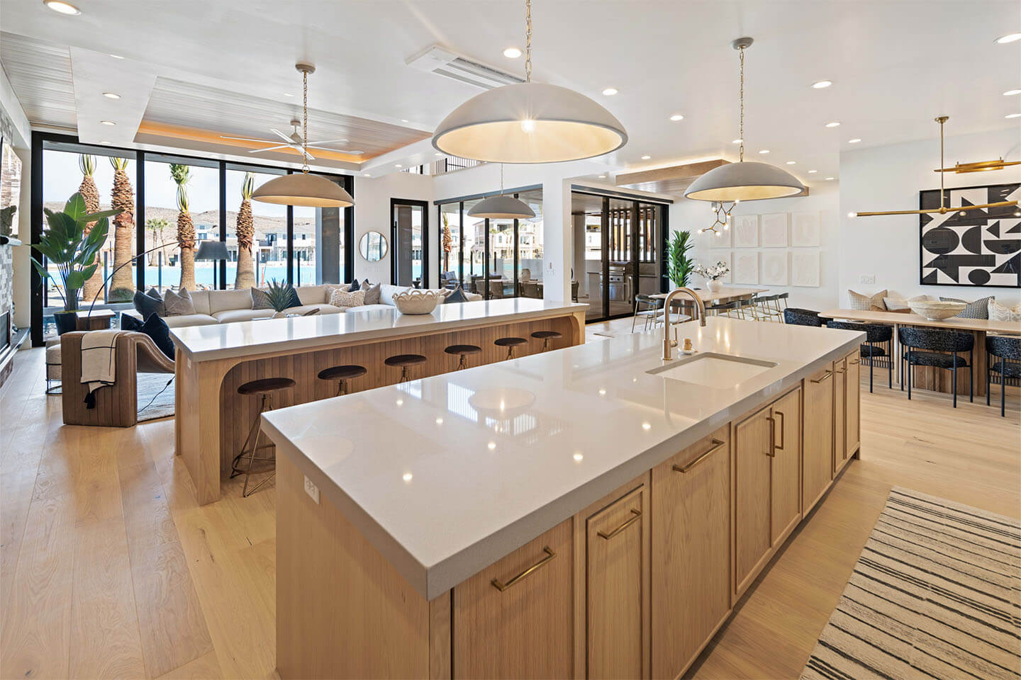 Spacious modern open kitchen with large white island countertops, wooden cabinetry, pendant lights, and adjacent living and dining areas with large windows.