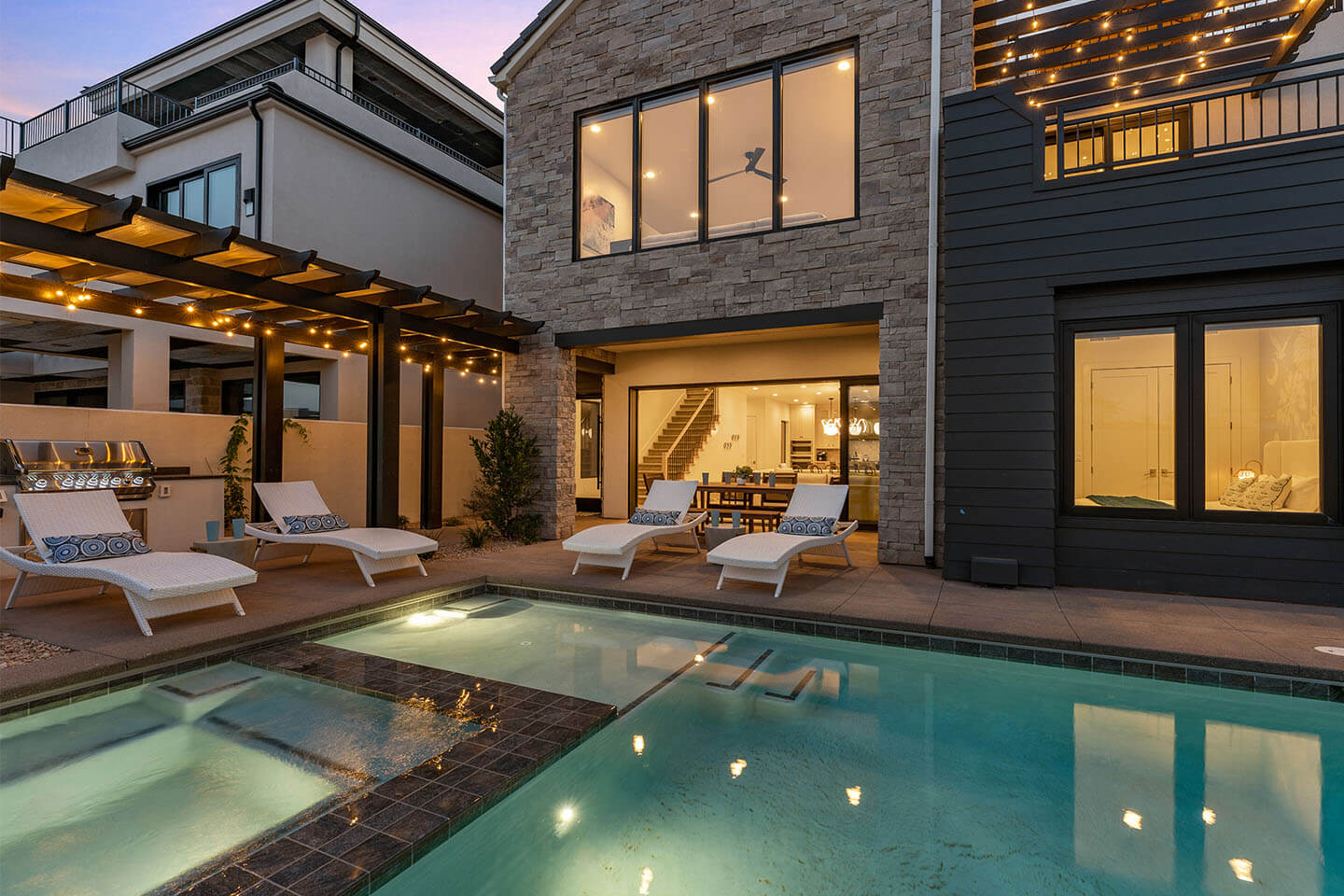Modern backyard with illuminated swimming pool, four white lounge chairs with blue patterned pillows, a barbecue grill, and a stone facade house with large windows and warm interior lights.