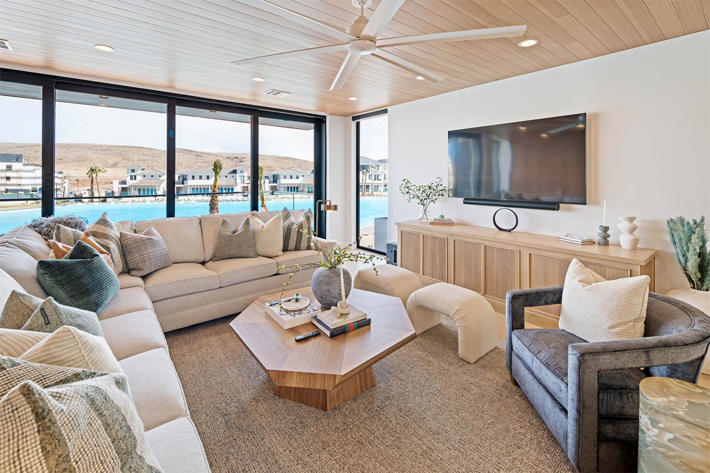 Modern living room with a large beige sectional sofa, wooden coffee table, TV mounted on the wall, and large windows overlooking a pool and hillside houses.