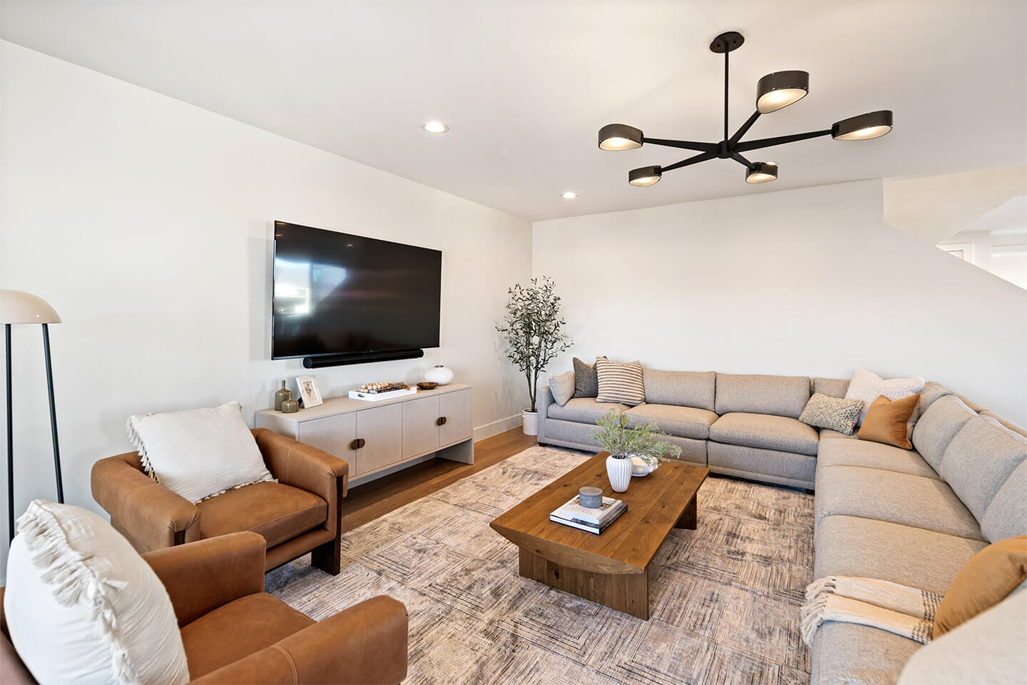 Modern living room with a large gray sectional sofa, two brown leather armchairs, a wooden coffee table, and a wall-mounted flat screen TV.