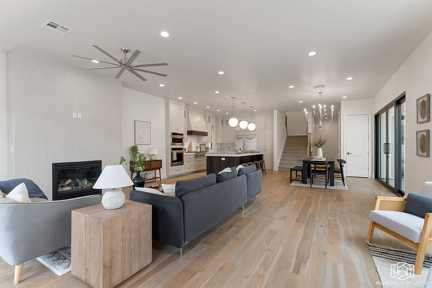 Modern open-concept living room with gray sofas, wooden floor, contemporary kitchen, and dining area with staircase in the background.