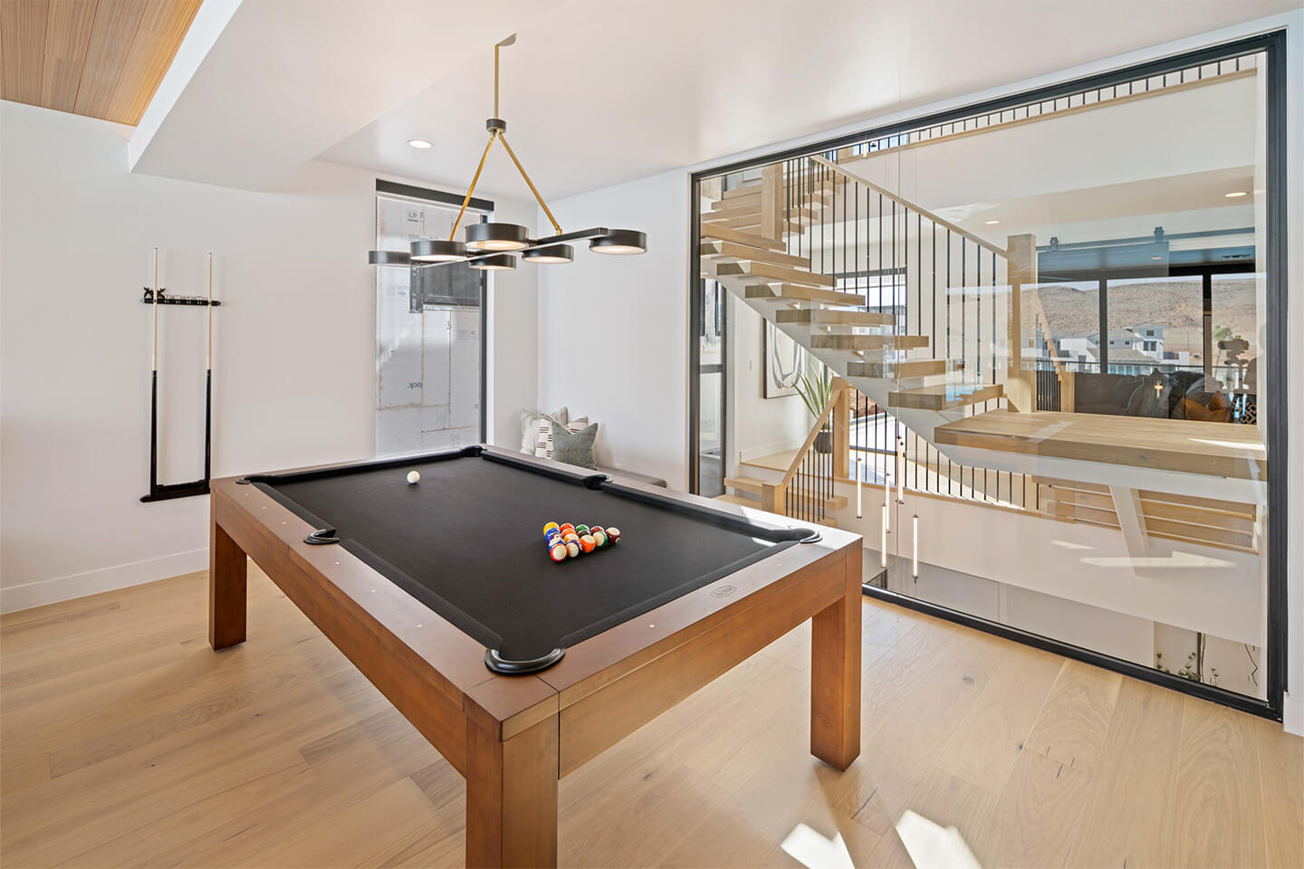Modern game room with a wooden pool table featuring black felt, a rack of pool cues on the wall, and a view of a floating staircase through a glass partition.
