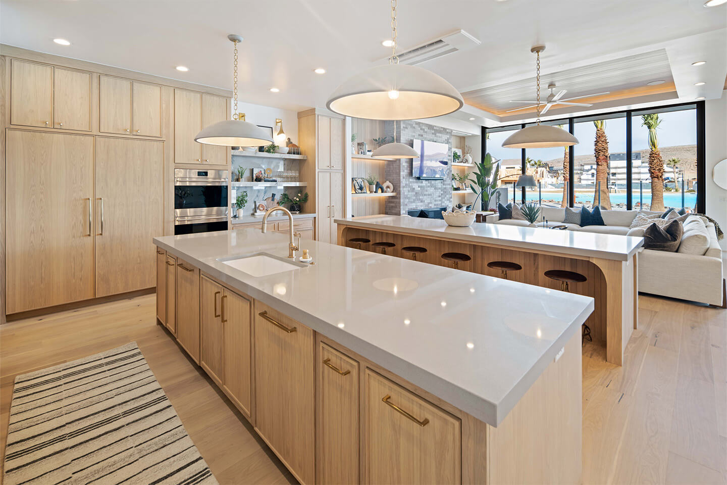 Modern open-concept kitchen with light wood cabinetry, two large islands with white countertops, pendant lights, and a living area featuring large windows with a view of palm trees and water.