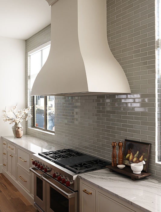 Modern kitchen with white curved range hood, gray subway tile backsplash, stainless gas range, marble countertops, and brass cabinet hardware