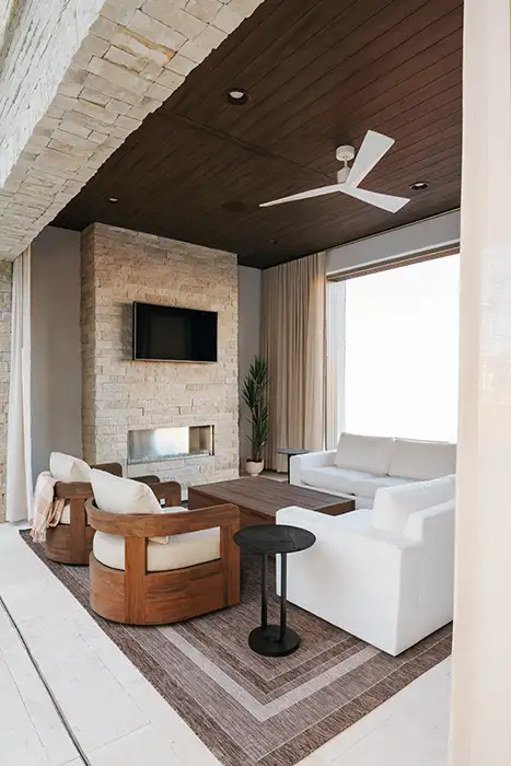 Covered outdoor living space with a stacked-stone fireplace, wall-mounted TV, wood plank ceiling, ceiling fan, and white upholstered sofas.