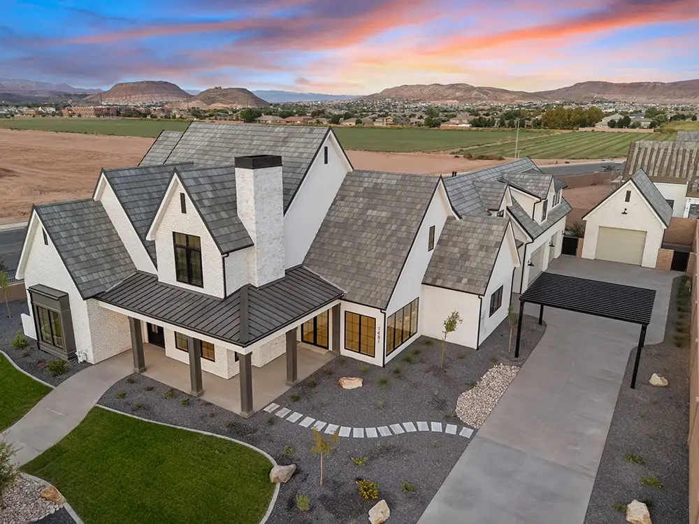 Aerial view of a white modern farmhouse with dark slate roof, chimney, and multi-car garage set against a desert sunset and mountain backdrop.