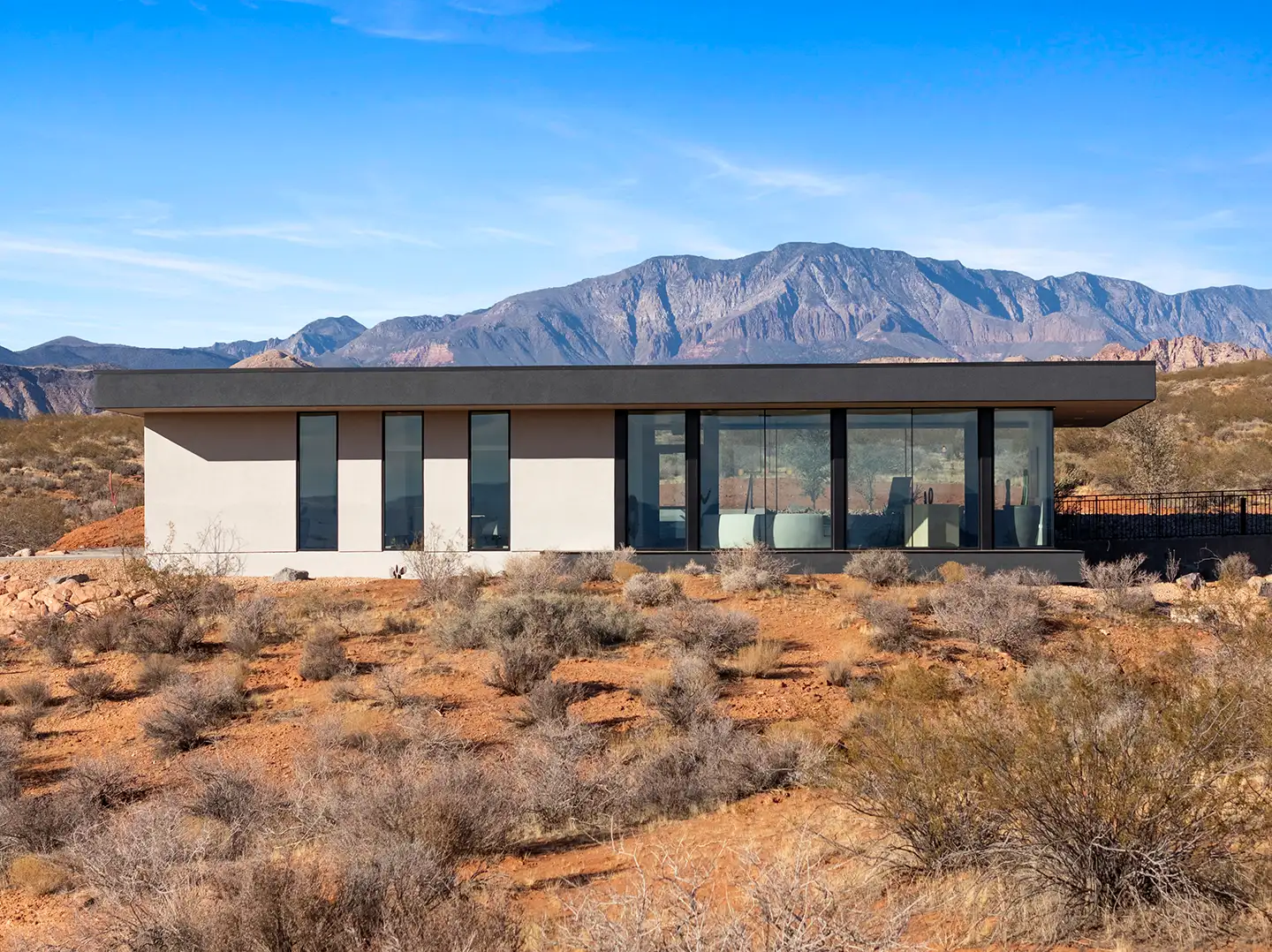 Modern single-story home with floor-to-ceiling glass walls on a desert hillside overlooking rugged mountain peaks.