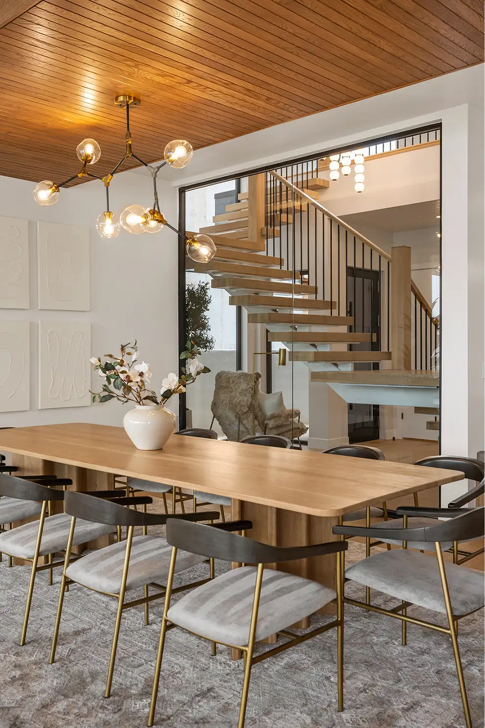 Modern dining room with live-edge oak table, upholstered chairs with brass legs, bubble chandelier, and open wood staircase behind glass partition