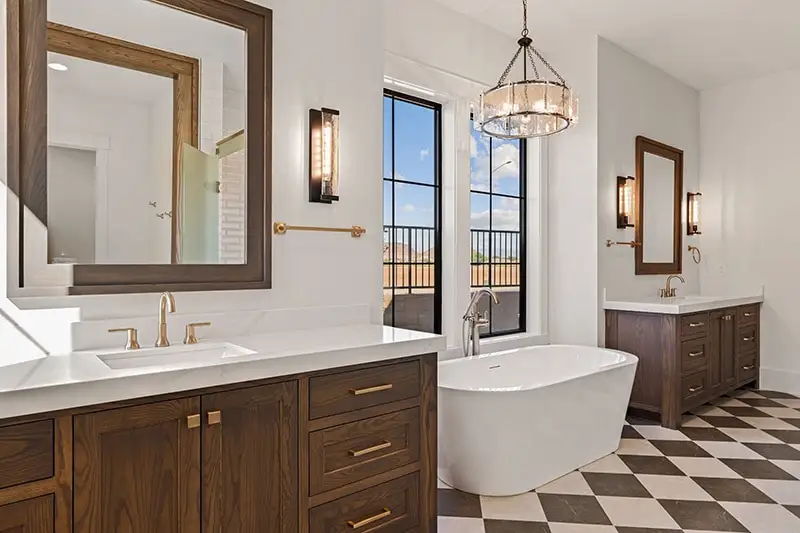Luxury primary bathroom with dual dark wood vanities, white freestanding soaking tub, checkered tile floor, and crystal chandelier