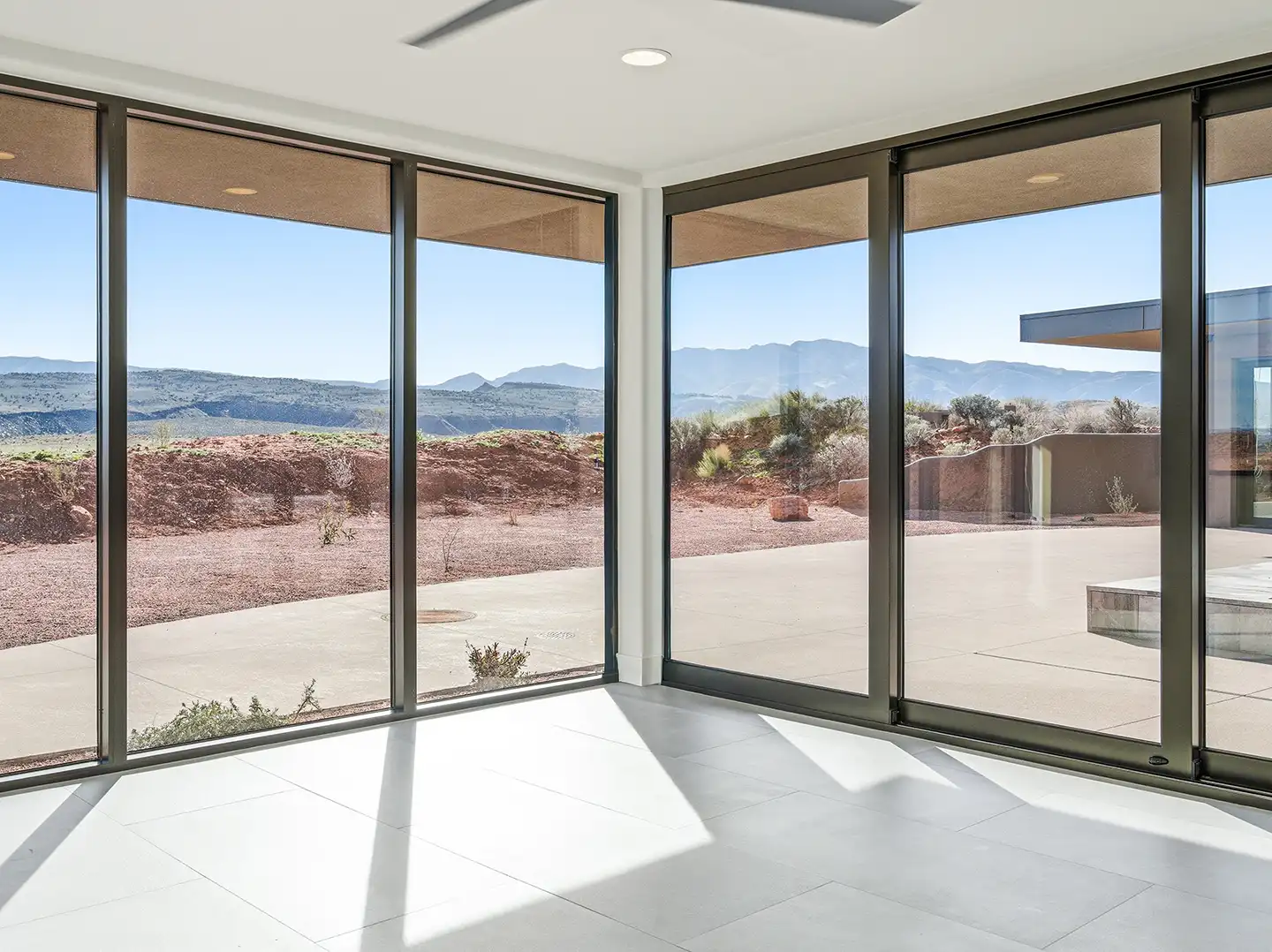 Corner room with floor-to-ceiling glass walls offering panoramic views of red desert hills and distant mountains.