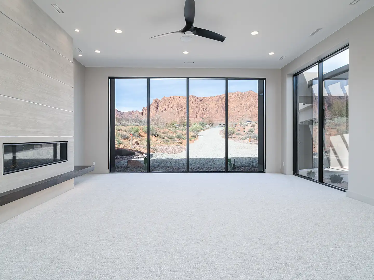 Open living room with linear fireplace, ceiling fan, and floor-to-ceiling sliding glass doors framing red rock cliffs.