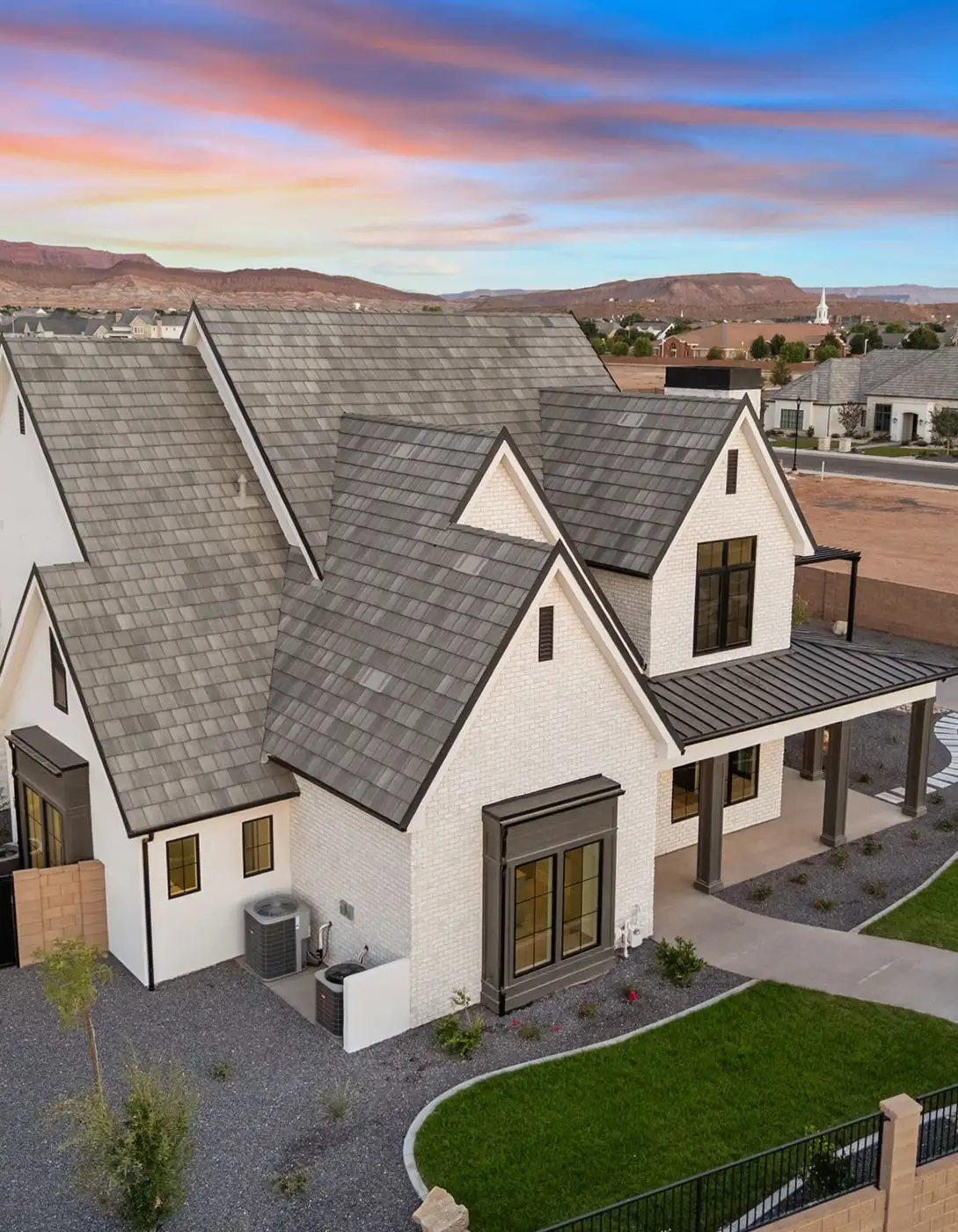 Aerial view of a white brick farmhouse with dark gray shingle roof and black windows against red rock mountains at sunset.