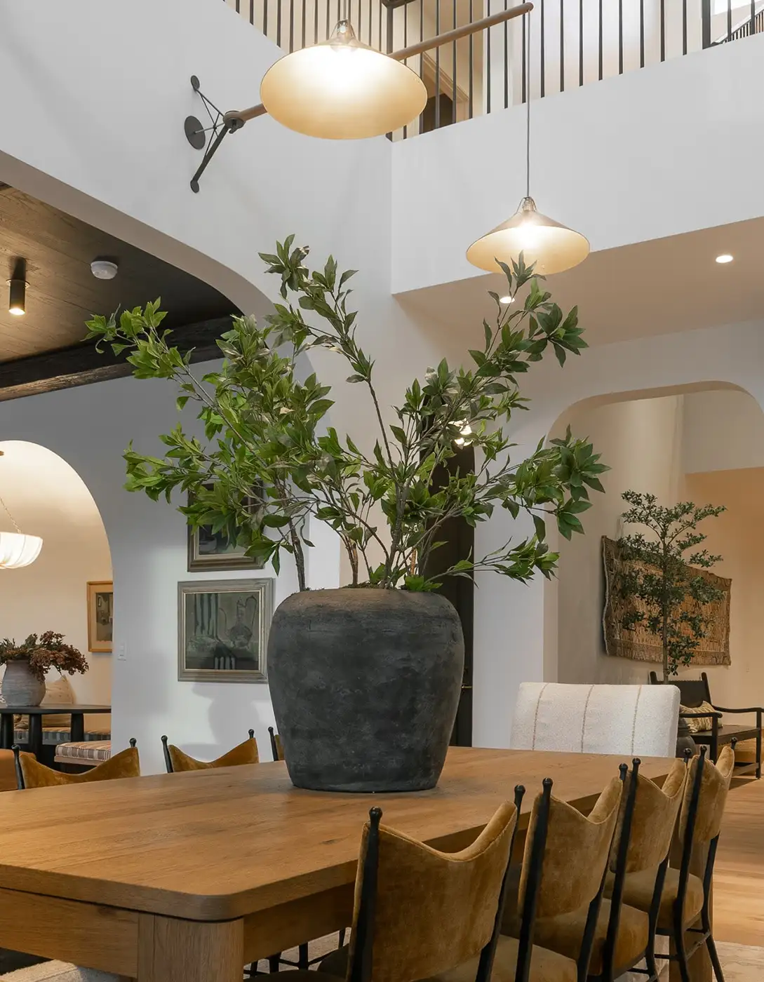 Open-concept dining room with wood table, leather chairs, an oversized black ceramic vase with tall greenery, and globe pendant lights.