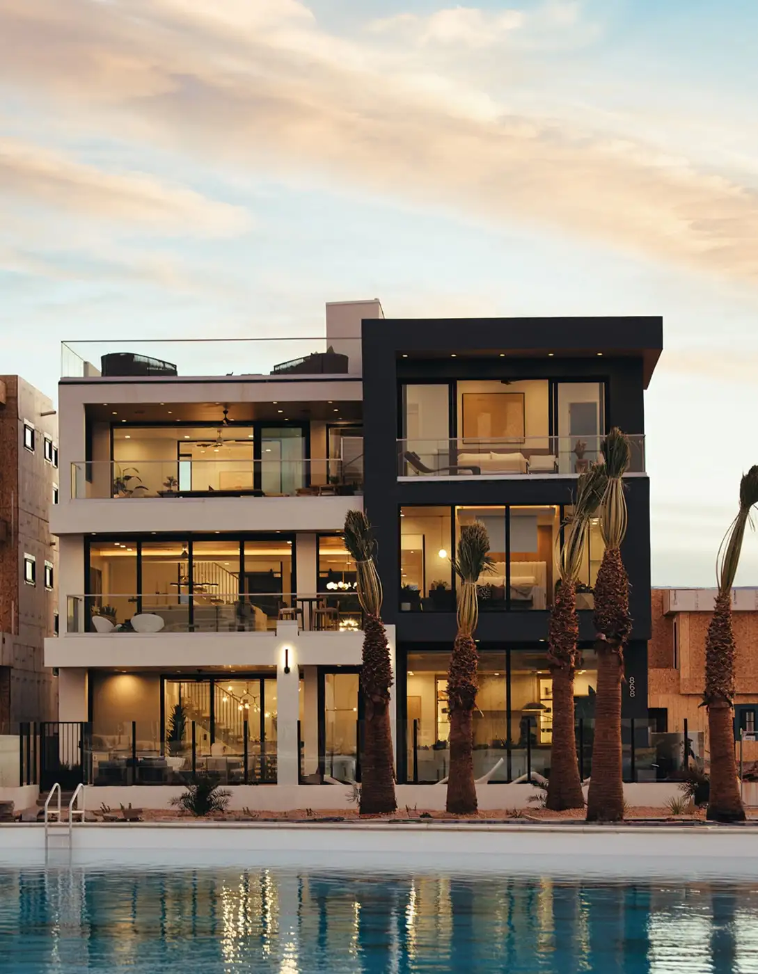 Modern two-story beachfront home at dusk with black-and-white facade, floor-to-ceiling glass walls, balconies, palm trees, and pool reflection.
