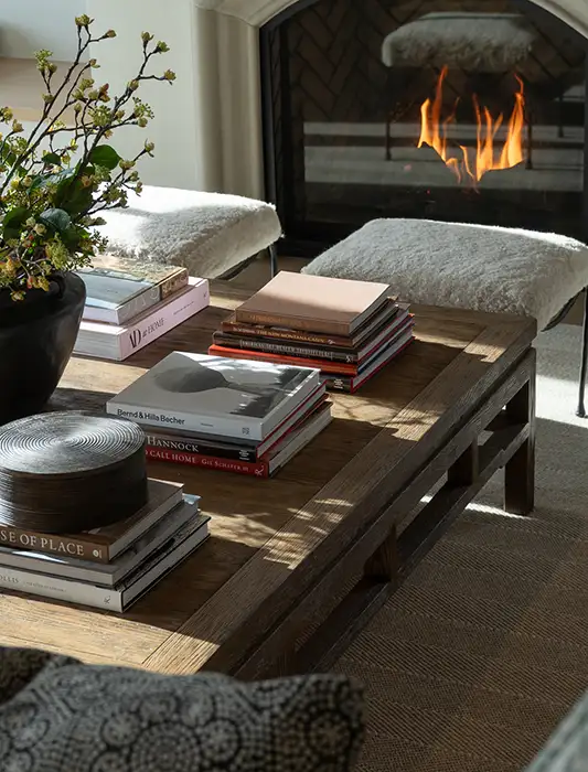 Styled wood coffee table with stacked design books and a plant beside a lit stone fireplace with sheepskin-covered benches