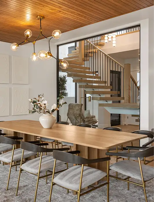 Modern dining room with oak table, brass bubble chandelier, wood-paneled ceiling, and a floating staircase visible through a glass wall