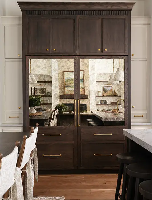 Floor-to-ceiling dark wood kitchen cabinet with antiqued mirrored glass panels and brass hardware flanked by light cabinetry