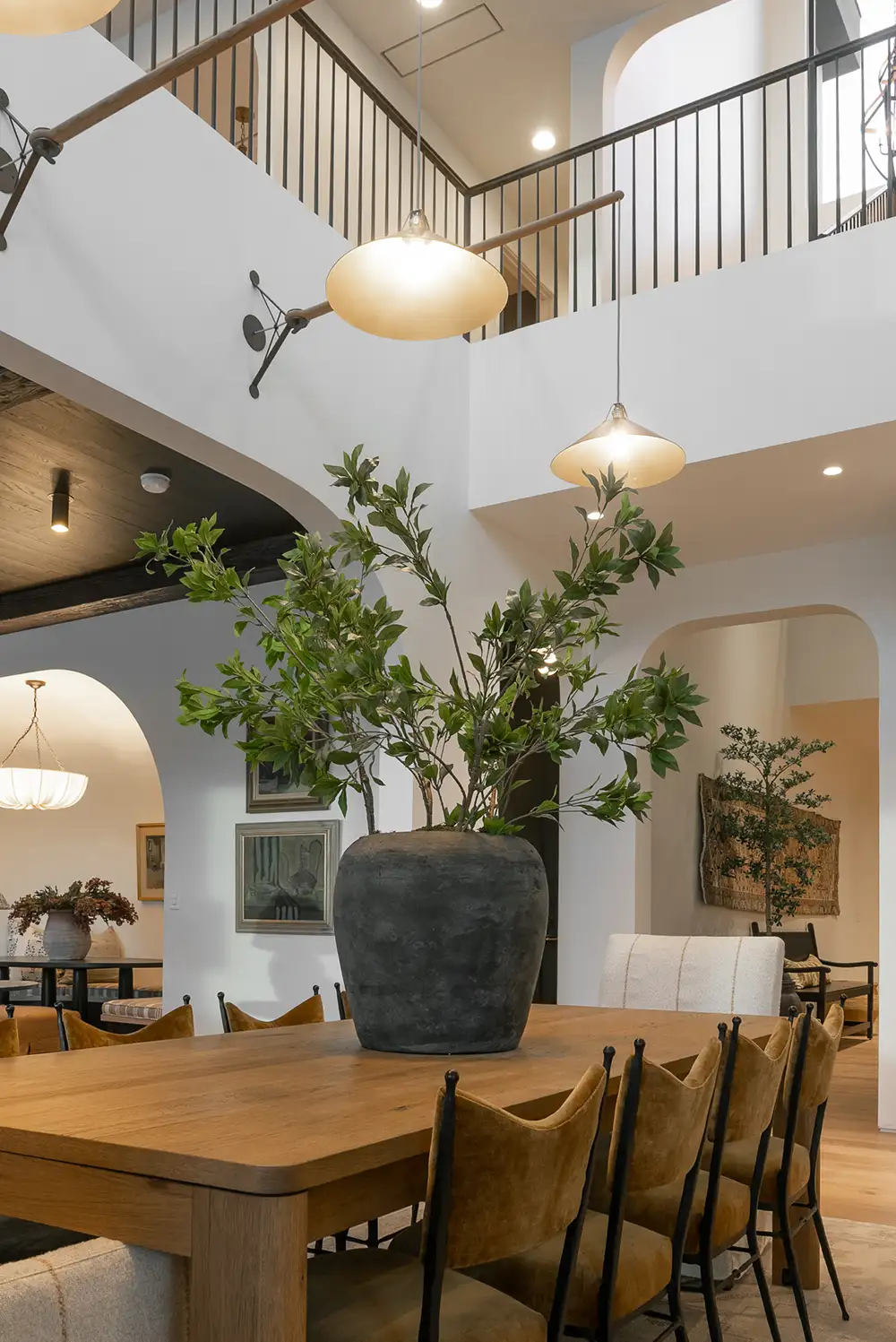Elegant two-story dining room with oak table, velvet chairs, pendant lighting, and a tall vase of fresh greenery.