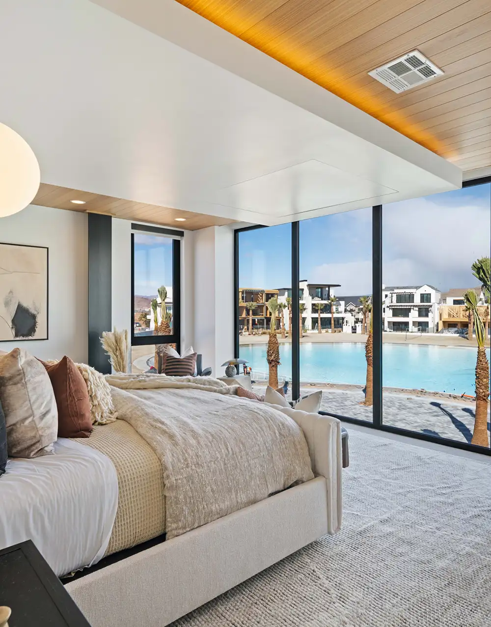 Bedroom with neutral bedding and floor-to-ceiling windows overlooking a lagoon with palm trees and modern homes across the water.
