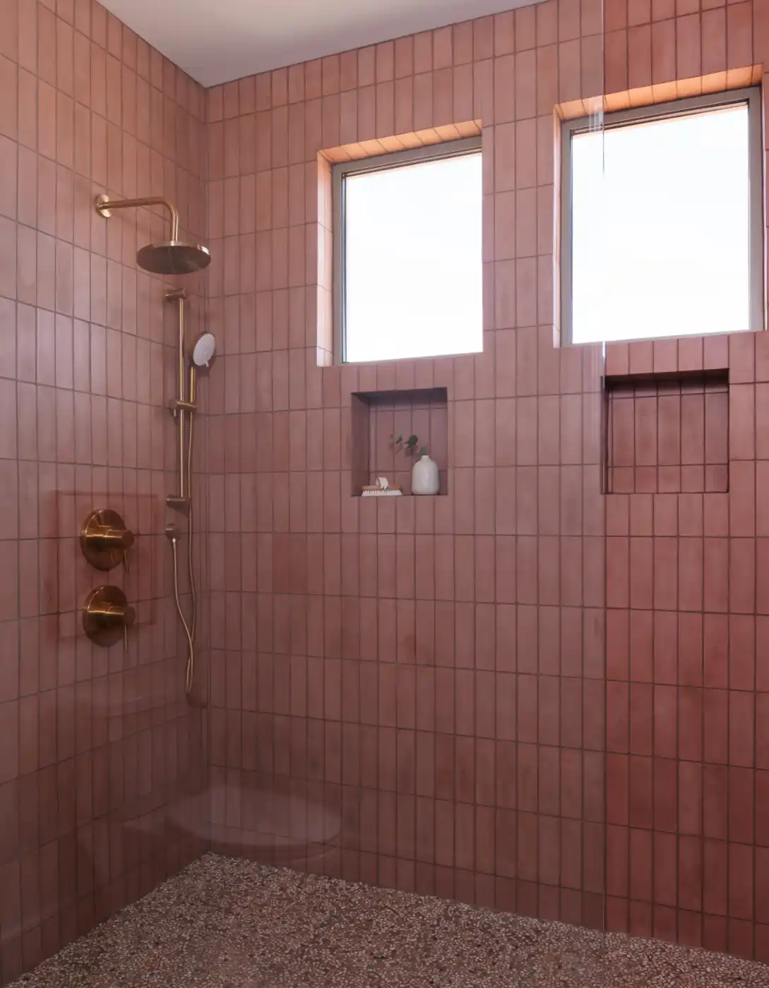 Modern shower with vertical pink terracotta tile walls, brass rainfall fixtures, recessed niches, and natural light from two upper windows