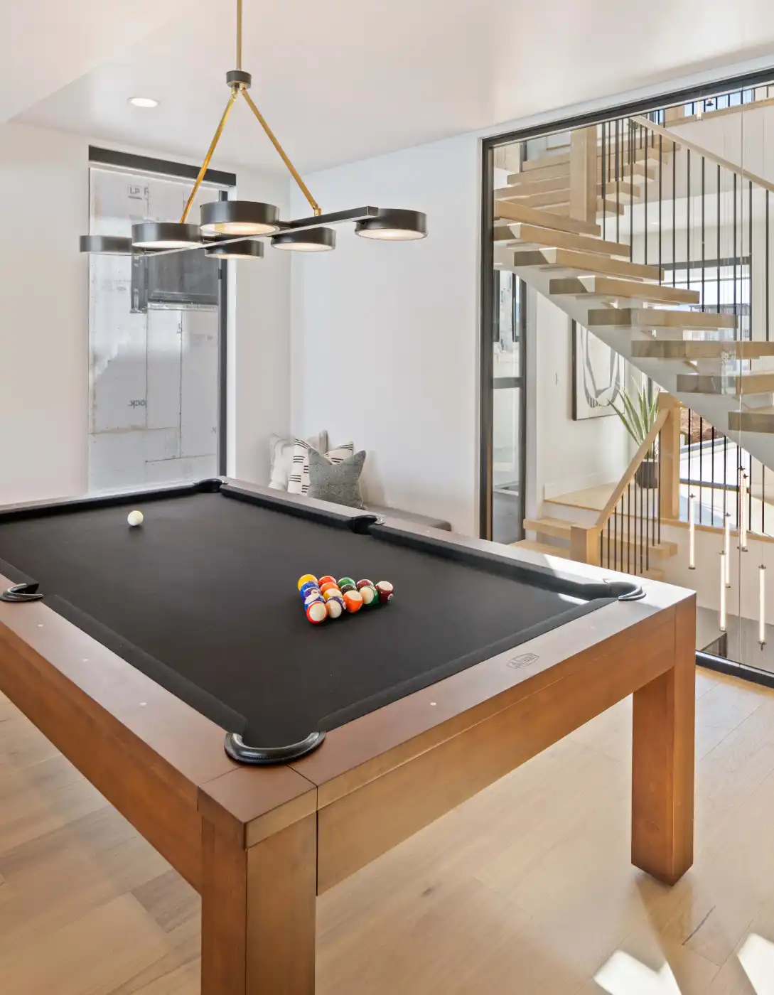 Modern game room with a wood-trimmed pool table, statement chandelier, light-wood floating staircase, and neutral living-area decor