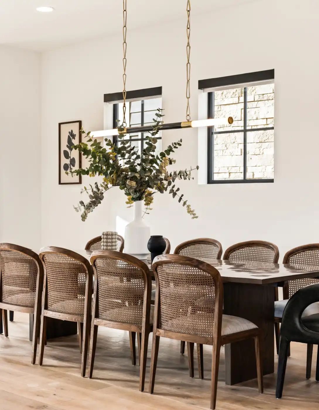 Bright dining room with a long wood table, cane-back chairs, a slim modern chandelier, and a tall eucalyptus arrangement in a white vase