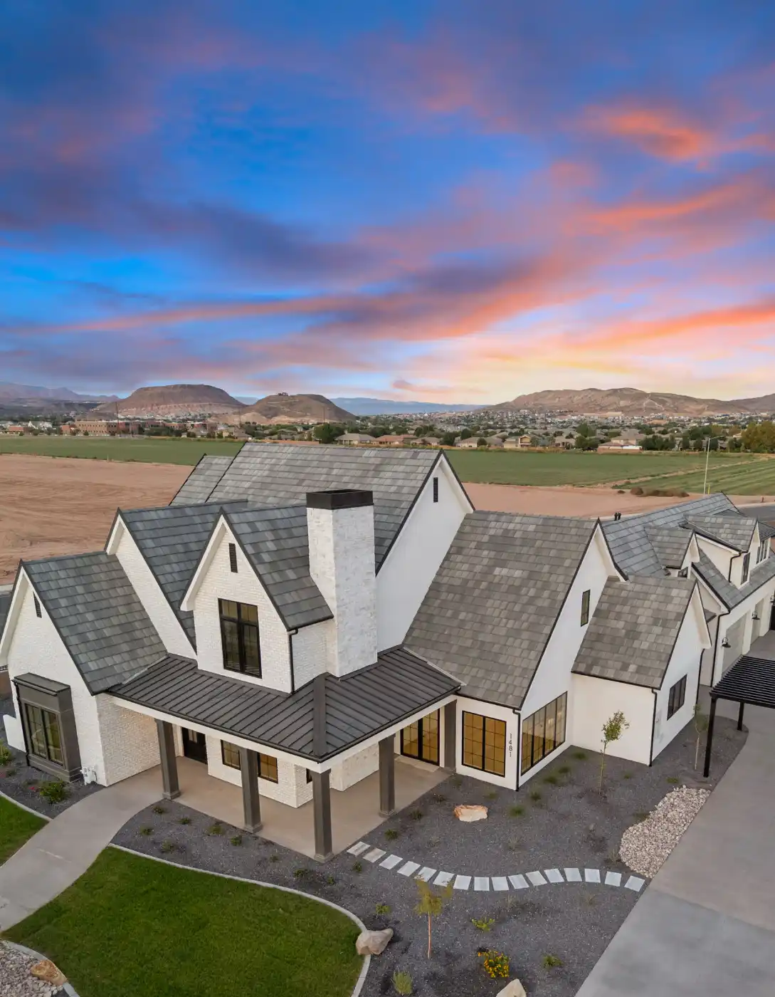 Aerial view of a modern farmhouse with white brick, slate roof, and a stone chimney at sunset with a vibrant pink and blue sky overhead