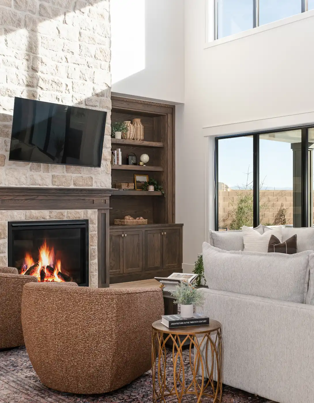 Living room with a stacked stone fireplace, mounted TV, wood built-in shelves, a neutral sofa, and boucle swivel chairs by a sunlit window
