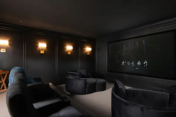 Dark home cinema room with plush sofas, illuminated wall sconces, panelled walls, and a large screen mounted on the back wall.