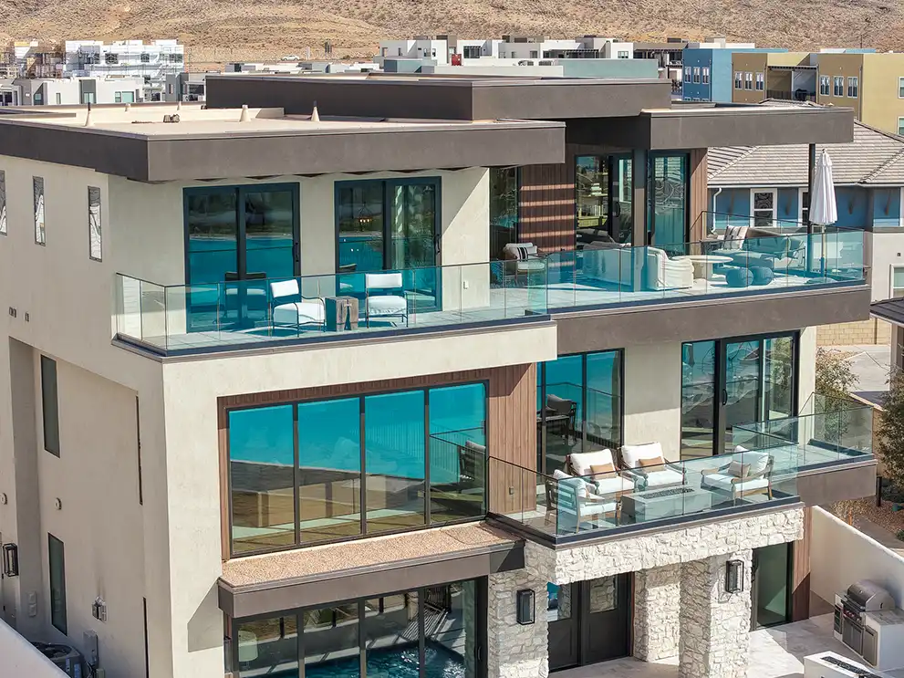 Modern luxury home exterior with large glass balconies furnished with outdoor seating overlooking nearby houses.