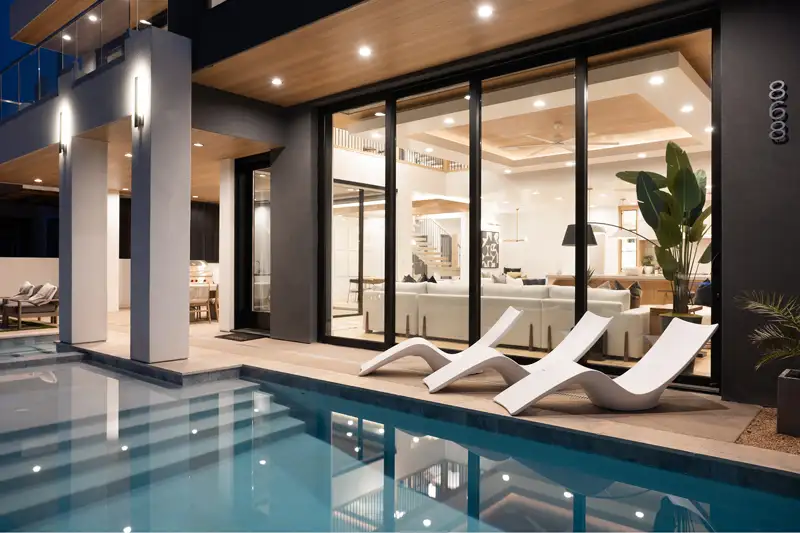 Modern home exterior at dusk with curved poolside lounge chairs, glass sliding doors, and a glowing interior living space.