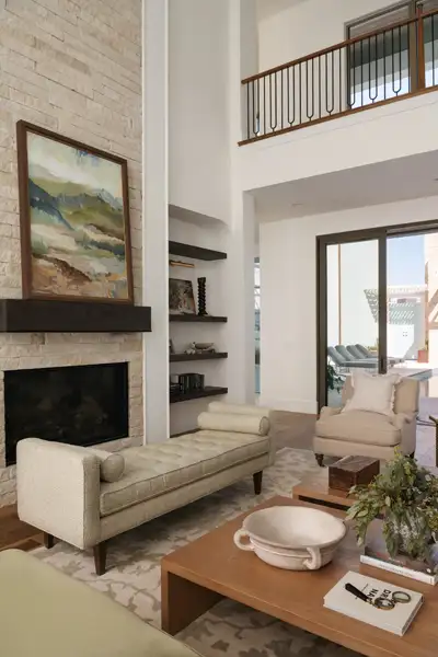 Two-story living room with brick fireplace, framed landscape art, upholstered bench, floating shelves, and a balcony railing above.