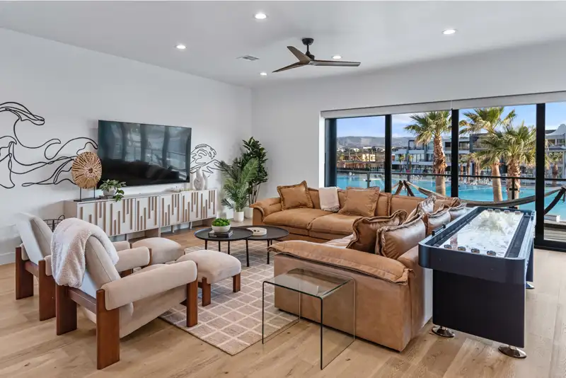 Living room with leather sofas, wall-mounted TV, shuffleboard table, and large windows overlooking a pool lined with palm trees.