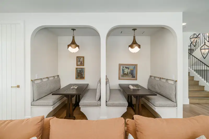 Breakfast nook with two arched alcoves, cushioned banquette seating, wood tables, pendant lights, and a staircase beyond.
