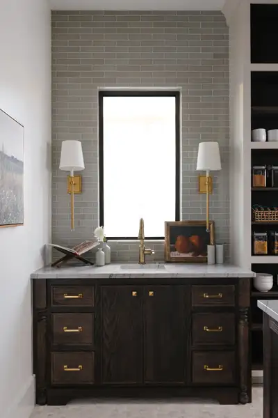 Wet bar with dark wood cabinetry, marble counter, gold faucet, green tile backsplash, two wall sconces, and a window above the sink.