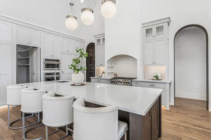 White kitchen with a large island, upholstered barstools, globe pendant lights, a range with hood, and arched doorways.