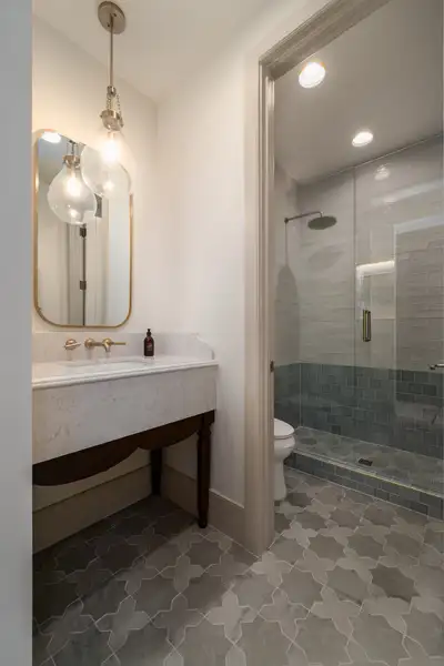 Bathroom with marble vanity, gold-framed mirror, pendant lights, and a glass walk-in shower beside a toilet on patterned tile floor.