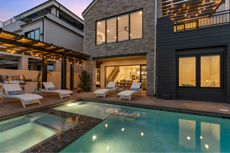 Backyard at dusk with a pool and spa, lounge chairs, outdoor grill, string lights, and a two-story stone home behind.