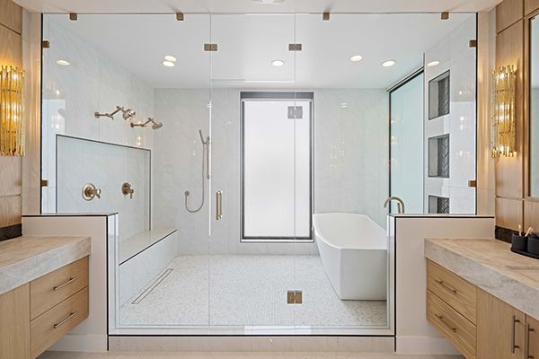 Bright primary bathroom with a glass-enclosed shower, freestanding white tub, dual wood vanities, and gold wall sconces.