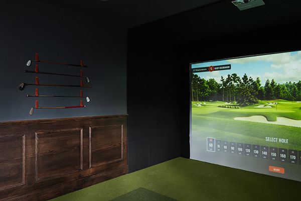 Indoor golf simulator room with clubs mounted on a dark wall, green turf floor, and a Trackman course on a large screen.