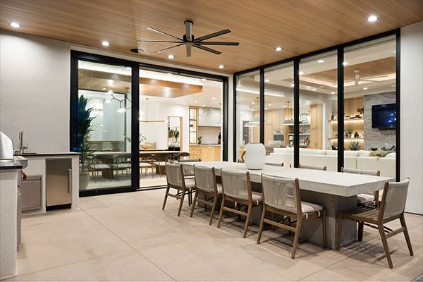 Covered patio with long concrete dining table, ceiling fan, and built-in grill, with sliding glass doors open to the interior.