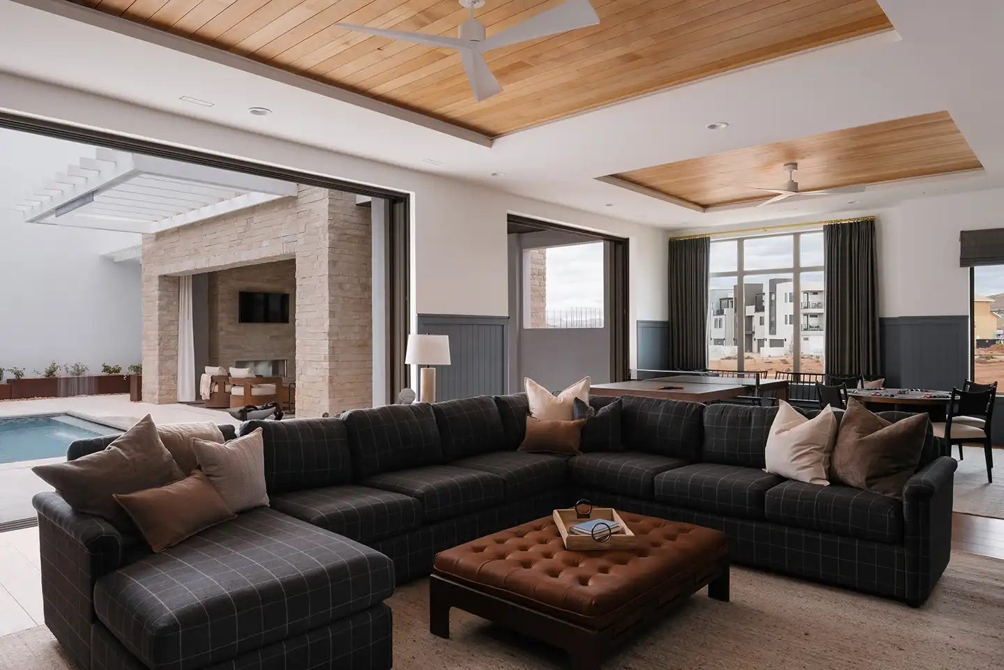 Modern open-concept living room with a large plaid sectional and leather ottoman, opening to a pool through glass walls.