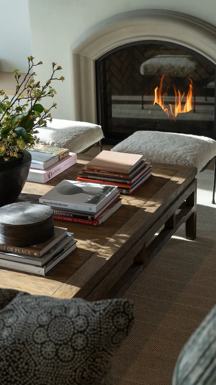 Sunlit living room with a lit arched fireplace, sheepskin benches, and a wood coffee table stacked with design books.