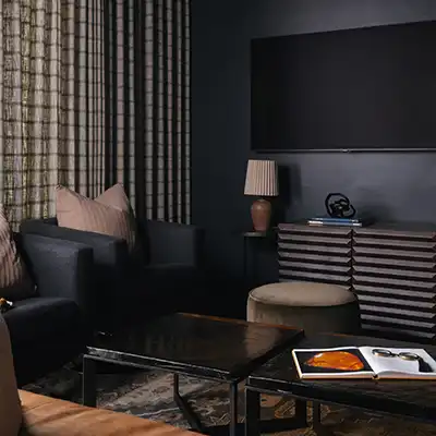 Dark, moody lounge with black walls, a wall-mounted TV, black armchairs, striped curtains, and a low coffee table.