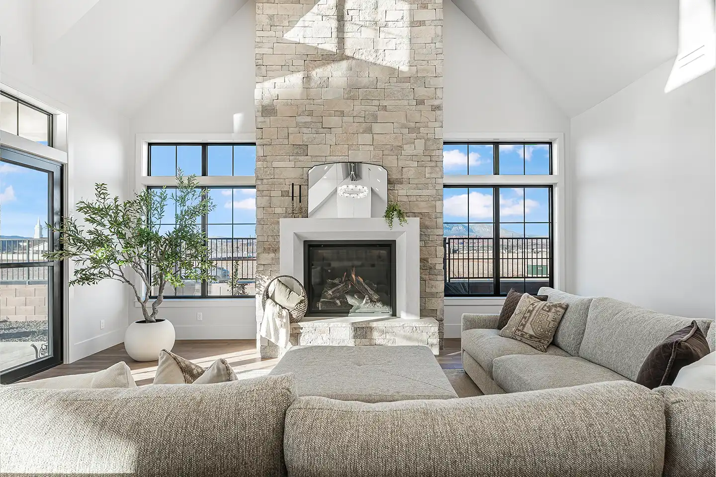 Bright living room with vaulted ceiling, stone fireplace, large windows, gray sectional sofa, and a potted plant.
