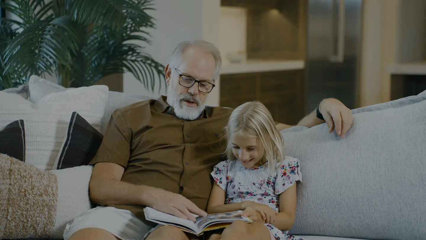 An older man in glasses reads a picture book with a young girl as they sit together on a sofa in a bright living room.