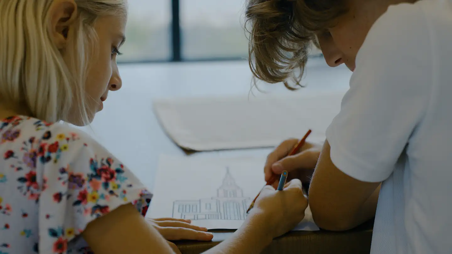 Two children lean over a sheet of paper at a table, drawing a large building with a central tower using colored pencils.