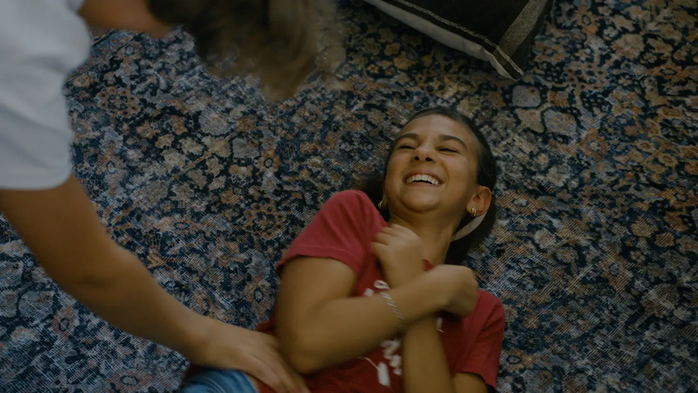 A girl in a red T-shirt lies on a patterned area rug, laughing, as another person reaches down to tickle her.