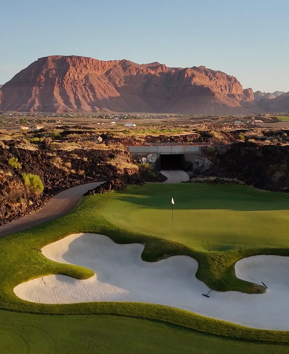 Manicured golf green with sculpted white sand bunkers and a cart tunnel framed by a red desert mesa at golden hour