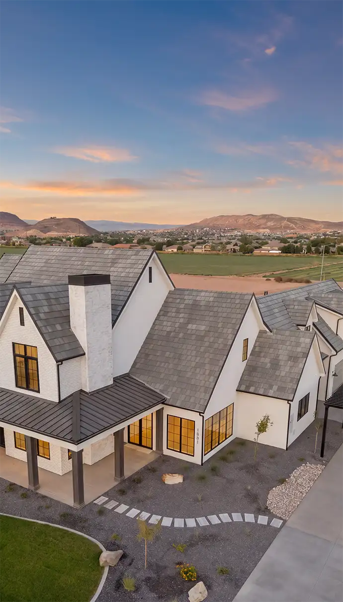 Aerial view of a white farmhouse-style home with a gray roof and mountains at sunset